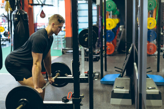 Fototapeta Gym-goer lifting loaded barbell at squat rack with punching bag, bumper plates, mirror reflections