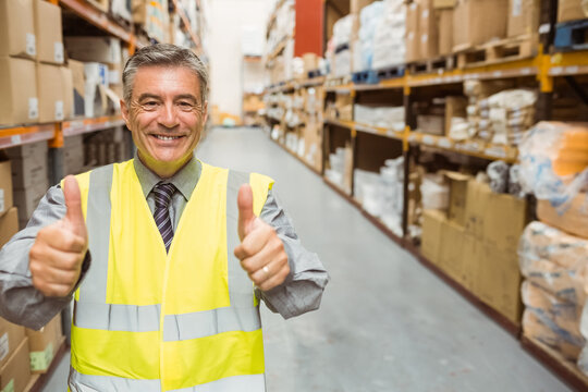 Male warehouse supervisor smiling wearing safety vest and giving thumbs up among shelving and boxes