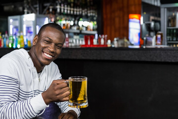 African American man wearing white hoodie sitting at pub counter holding clear beer mug, copy space