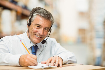 Male in business attire writing on clipboard with headset leaning on box in warehouse, copy space