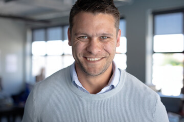 Obraz premium Man in his 30s wearing light-blue sweater smiling at camera in office with desks, large windows