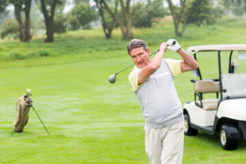 Senior man swinging driver golf club on fairway in polo shirt near golf cart, copy space