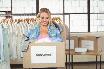 Female volunteer holding donation box sorting clothes in donation center near rack and hatbox