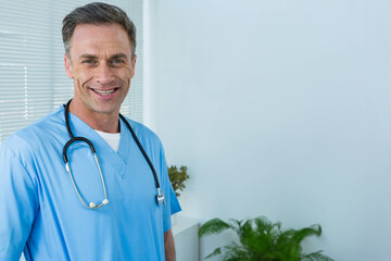 Male doctor standing and smiling at viewer in clinic with blue scrubs, stethoscope, copy space