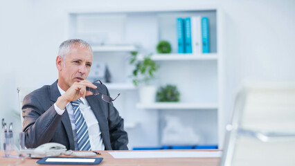 Fototapeta premium Senior man wearing business suit sitting at desk in office holding eyeglasses, copy space