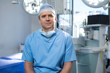 Male surgeon sitting in surgery room in scrubs beside anesthesia machine under overhead lights