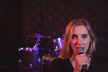 Female singer singing into microphone on stage under purple lights with drum kit, copy space