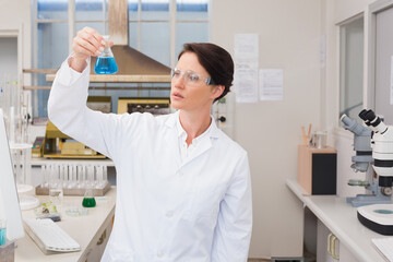 Female scientist holding up conical flask in lab inspecting blue solution wearing goggles