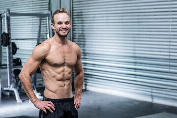 Obraz premium Shirtless muscular man smiling at camera in gym with weight machine, metal shutter door, copy space