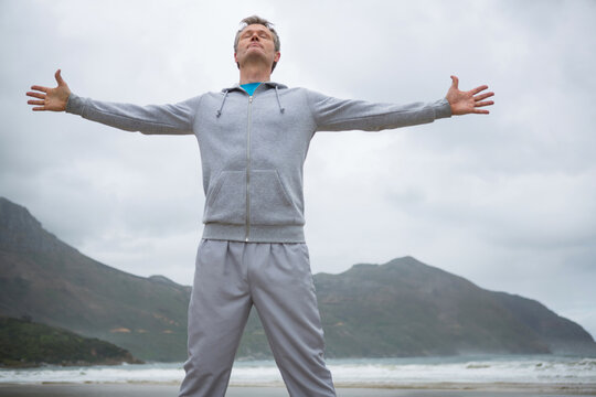 Fototapeta Middle-aged man in grey hoodie and sweatpants stretching arms and inhaling ocean breeze on beach