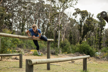 Woman vaulting over wooden beam in clearing on obstacle course wearing blue top, black leggings
