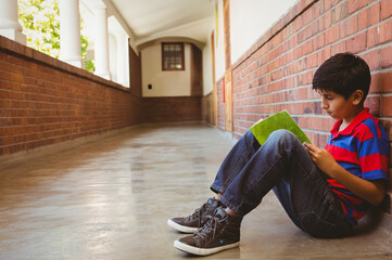 Boy sitting in brick corridor reading green book near white columns with greenery beyond