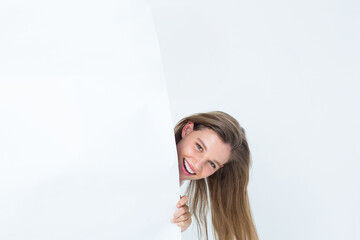 Female model peeking behind large blank panel at studio, smiling and gripping edge, copy space