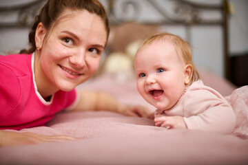 Young woman with brown hair and smiling baby girl on pink bed. Light-hearted interaction, creating joyful atmosphere. Warm lighting enhances bond