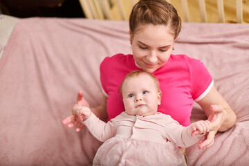 Young mother with brown hair cradles baby girl on cozy pink couch. Warm lighting highlights their...