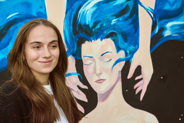 Young adult female with brown hair stands beside colorful canvas. Mural shows stylized face with...