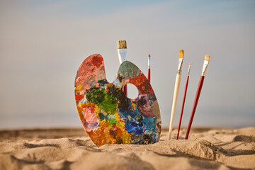 Vibrant painter palette and brushes rest on tranquil beach, symbolizing creativity and inspiration....