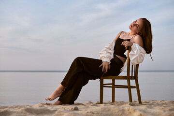 Fototapeta premium Young female painter sits thoughtfully on chair by tranquil beach. Serene setting and soft lighting evoke creativity and introspection, symbolizing artistic inspiration