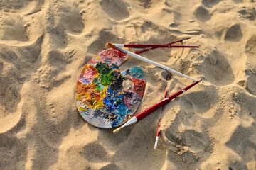 Artist palette with vibrant paints lies on sunlit sand, surrounded by brushes. Scene evokes...