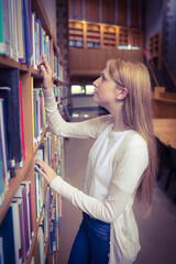 Female student is browsing wooden bookshelves and reaching for book in library hall under warm glow