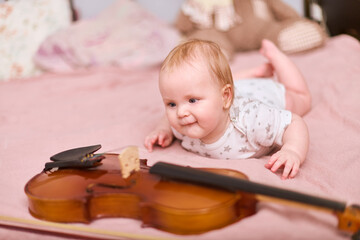 Baby girl, blonde, caucasian, lies on bed observing violin closely. Soft lighting, warm ambiance....