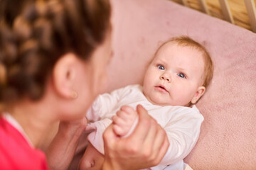 Caucasian baby girl with light brown hair happily lying on soft pink blanket. Adult single mother gently holding baby hands. Soft lighting creates warm atmosphere