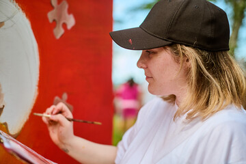 Young caucasian female painting vibrant mural with brush, daylight illuminating artwork. Red,...