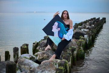 Young female artist peacefully holds her painting while sitting on coastal rocks. Tranquil seascape...