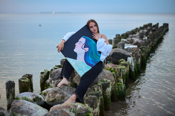 Young female artist peacefully holds her painting while sitting on coastal rocks. Tranquil seascape...