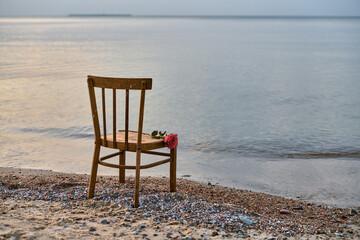 Empty wooden chair sits on serene beach, accompanied by single rose, against sunset backdrop. Image evokes feelings of solitude, reflection, and peace