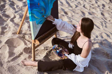 Young female artist works on vibrant painting, seated on sandy beach. Serene coastal environment...