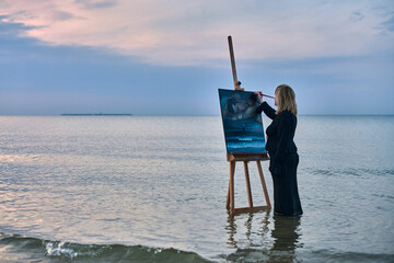Young woman stands in shallow water, painting serene ocean scene on easel at dusk. Tranquil...