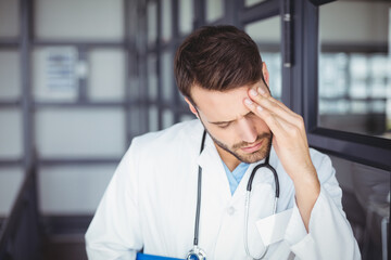 Male doctor leaning against glass cabinets in medical corridor, holding blue folder and stethoscope