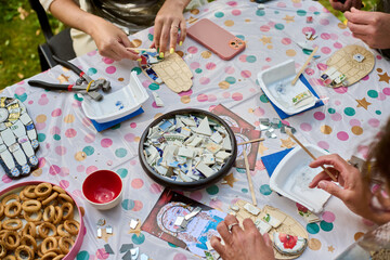 Hands crafting mosaic on polka dot tablecloth outdoors. Bright sunlight highlights ceramic pieces,...