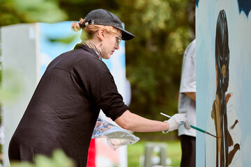Young female artist with cap and gloves painting on large canvas in sunlight. Surrounded by...