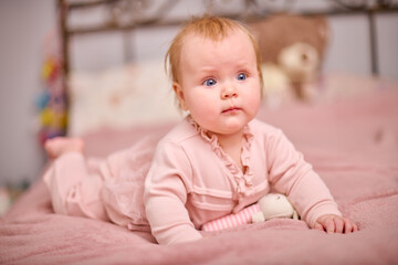 Fototapeta premium Baby girl with light hair and blue eyes lying on pink bed. Soft lighting and bedroom setting create warm and peaceful atmosphere