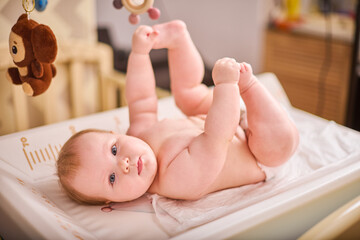 Young caucasian infant with light brown hair lies on changing table engaging with hanging toys. Soft lighting and neutral background create calm atmosphere