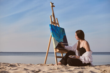 Young female artist paints on beach, capturing serene ocean view. Setting evokes tranquility and...