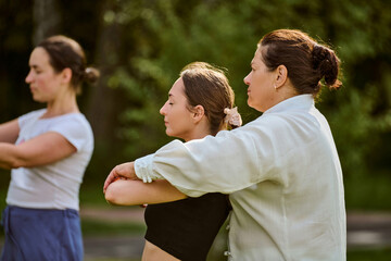 Mature woman instructing young woman in qigong outdoors. Olive-skinned females focus on balance and energy flow. Sunlight filters through trees, creating serene atmosphere