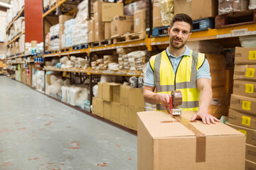 Male warehouse worker sealing box with tape dispenser in warehouse aisle with racks, copy space