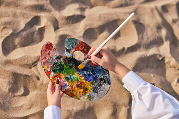 Young adult female artist uses paintbrush on colorful palette while seated on sunlit sandy beach....