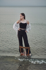 Young woman stands on submerged chair in shallow water, exuding confidence and poise. Her casual...
