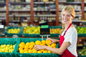 Female store employee in red apron arranging oranges in green bins at produce section, copy space