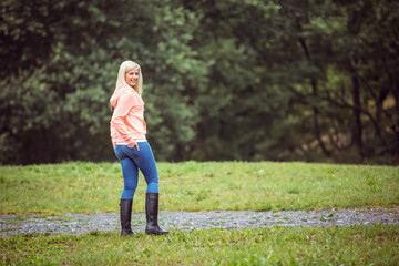 woman wearing pink hoodie, jeans, black rubber boots standing on gravel path among grass near trees