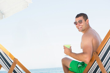 Man in swim trunks relaxing on chair under umbrella holding martini glass on beach, copy space