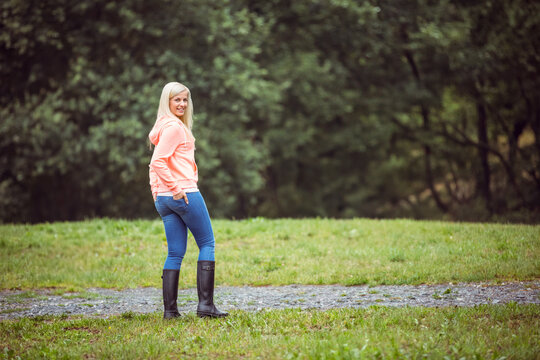 Fototapeta woman wearing pink hoodie, jeans, black rubber boots standing on gravel path among grass near trees