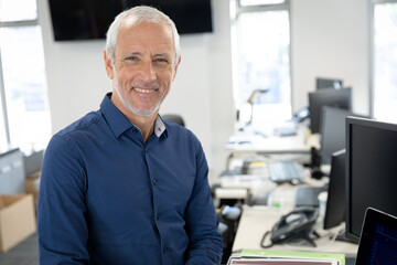 Senior man wearing blue shirt leaning against desk smiling in office with laptop, copy space