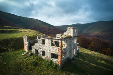 Abandoned stone building on grassy hillside, partially in ruins. Structure against backdrop of rolling hills and dense forest, under sky with dramatic clouds, evoking sense of history and solitude.