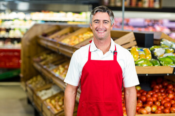 Smiling male grocery store employee wearing red apron standing in produce aisle with green peppers