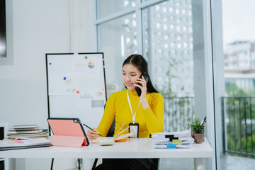 Image of young Asian woman, office worker busy with documents and smiling while holding a phone, office background.